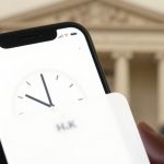 A smartphone displaying a clock app is held in the foreground. A classical building with columns is blurred in the background, along with a person standing with hands together.