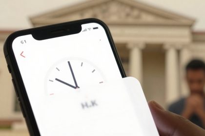 A smartphone displaying a clock app is held in the foreground. A classical building with columns is blurred in the background, along with a person standing with hands together.