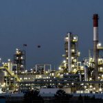 Industrial refinery at dusk with illuminated towers, pipes, and smokestacks. American and Chinese flags are visible against the evening sky.