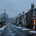 Snow-covered residential street with brick houses on either side. Two people sit on a sofa outside, lit by candlelight. Overcast sky.