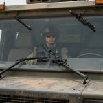 A person wearing a helmet and sunglasses sits inside a military vehicle, holding a rifle. The vehicle is on a road in a desert area.