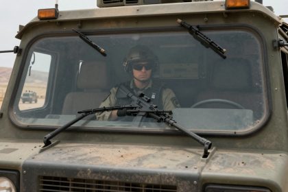 A person wearing a helmet and sunglasses sits inside a military vehicle, holding a rifle. The vehicle is on a road in a desert area.