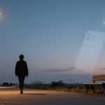 A person walks on a path at dusk under streetlights, with a bench nearby. A crescent moon is visible in the clear sky.