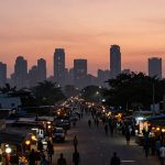 A street market scene at dusk with people walking and shopping. Stalls with colorful lights line both sides, and tall city buildings are silhouetted against an orange sky in the background.