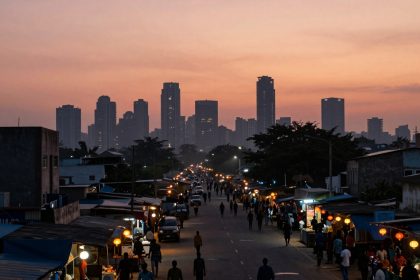 A street market scene at dusk with people walking and shopping. Stalls with colorful lights line both sides, and tall city buildings are silhouetted against an orange sky in the background.
