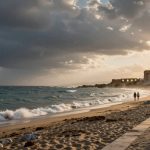 Sandy beach at sunset with waves crashing, people walking, and ancient ruins in the distance. Dark clouds cover part of the sky.