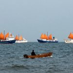 A person in a small wooden boat on a calm sea, with five larger ships in the background. The ships have orange sails and are painted white and blue.