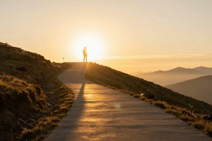 A person stands on a winding path atop a hill during sunrise, casting a long shadow. The sky is golden, and mountains are visible in the background.