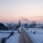 Snow-covered rural road at sunrise, bordered by houses with snowy roofs. A person shovels snow, and children walk in the distance. Smoke rises in the sky.