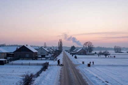 Snow-covered rural road at sunrise, bordered by houses with snowy roofs. A person shovels snow, and children walk in the distance. Smoke rises in the sky.