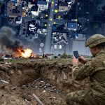 A soldier in camouflage and helmet uses a smartphone near a crater. Smoke and fire rise from burning buildings in the background. Overhead, a map of a city with highlighted points is visible. Debris is scattered around the scene.