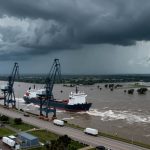Aerial view of a large cargo ship navigating a flooded river, with cranes along the dock. Dark storm clouds loom overhead, and trucks are on the nearby road.