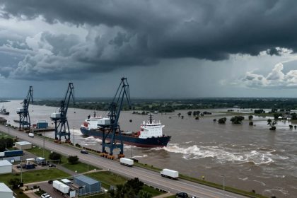 Aerial view of a large cargo ship navigating a flooded river, with cranes along the dock. Dark storm clouds loom overhead, and trucks are on the nearby road.