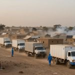 A convoy of white trucks with blue logos drives through a dusty village with mud brick houses and tents. People in blue uniforms walk alongside.