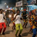 People dancing on a city street at night, some wearing colorful patterned clothing. Others hold smartphones. Motorbikes and tuk-tuks are visible.