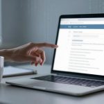 A hand points at a laptop screen showing a webpage with a red alert icon. A steaming cup is on the desk beside a notebook.