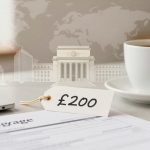 A mortgage document and a laptop are on a table. A tag labeled "£200" is placed on the document. A cup of coffee sits nearby. In the background, a faded image of classical buildings and a world map are visible.