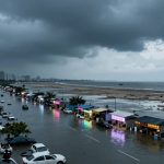 Flooded coastal street lined with cars and lit stalls. Overcast sky and distant city skyline. Beach and ocean visible to the right.