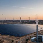 Aerial view of a large solar panel array and multiple wind turbines in a desert landscape at sunset. A plane flies overhead, and a factory with a smoking chimney is visible in the foreground. Hills are in the background.
