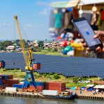 A large hand holds a smartphone displaying a mobile app, set against a background of a port with cranes, shipping containers, and solar panels. A market scene with blurred people and colorful stalls is partially visible.