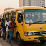 A group of people, some holding phones, stand and laugh near a yellow bus on a city street. Buildings and other vehicles are in the background.