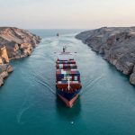 A large cargo ship navigates a narrow, rocky canal, carrying colorful shipping containers. Steep cliffs line the waterway under a clear sky.