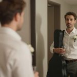 A man with a beard stands in front of a mirror, wearing a white shirt and holding a guitar case and a glass. A wedding photo is visible on the wall.