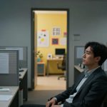 A man in a suit sits pensively in an office cubicle, surrounded by paperwork and a computer. The background shows a yellow-walled office with colorful art.