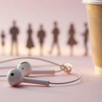 White earphones on a pink surface next to a brown paper cup. Blurred silhouettes of people are visible in the background.