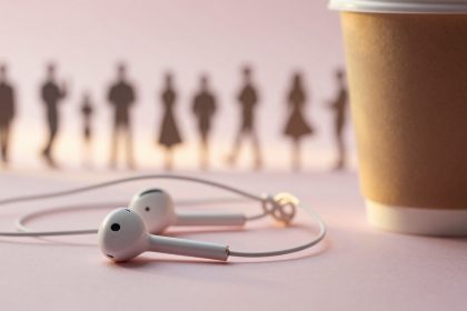 White earphones on a pink surface next to a brown paper cup. Blurred silhouettes of people are visible in the background.