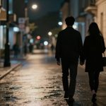 A couple walks hand in hand down a wet, dimly lit city street at night, surrounded by tall buildings and streetlights.