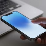 A hand holding a smartphone with a blue gradient screen, positioned above a silver laptop keyboard on a gray surface.