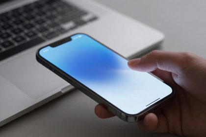 A hand holding a smartphone with a blue gradient screen, positioned above a silver laptop keyboard on a gray surface.