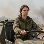 A woman in a military jacket drives a vehicle through a dusty, rubble-strewn area. Smoke and fire are visible in the background.