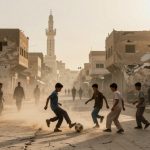 Five boys play soccer on a dusty street lined with damaged buildings and market stalls. A minaret is visible in the background.