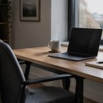 An empty chair at a tidy desk beside a laptop with a dimmed screen, soft daylight through a window